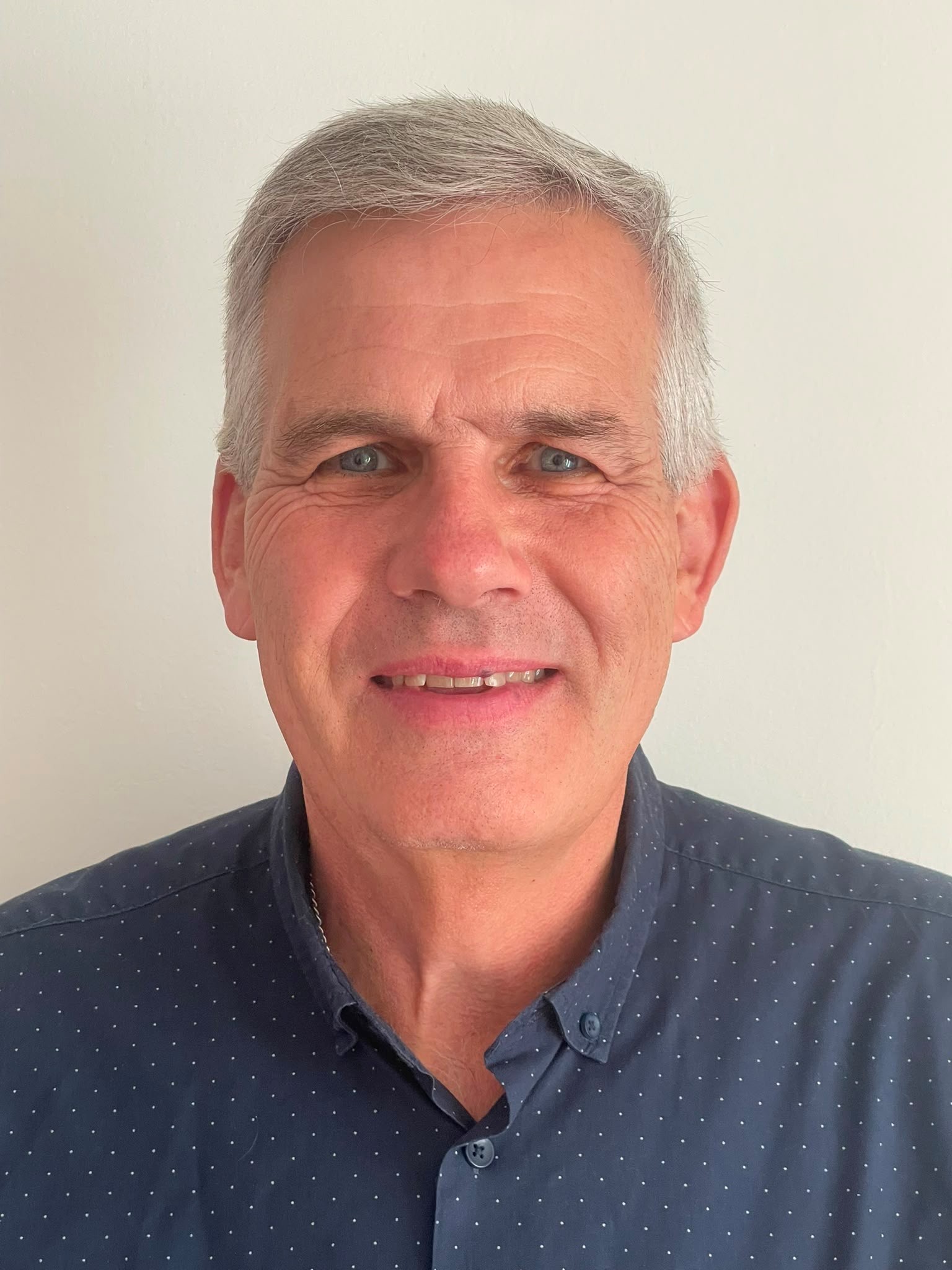 Philip is smiling. He is wearing a greyish blue shirt with white dots.