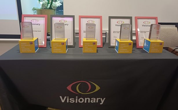 A table with a line of trophies sitting on top of a CreaCube unit and behind are certificates. The table has a tablecloth which has the Visionary logo on it.