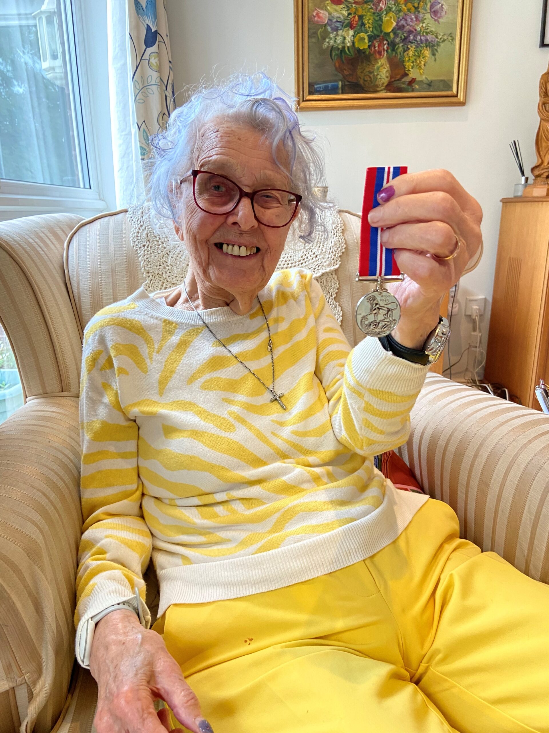 Gwenneth Pedler is sitting on a sofa, holding a medal.