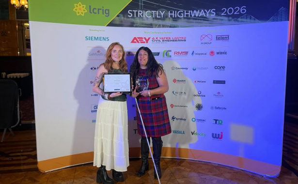 Seema Flower, founder of Blind Ambition, proudly receives the Outstanding Contribution Award (Private Sector) at the Strictly Highways 2026 awards ceremony. Seema, wearing a red and navy tartan dress and holding her white cane, stands alongside the award presenter, who holds a framed certificate, in front of the event’s sponsor backdrop.