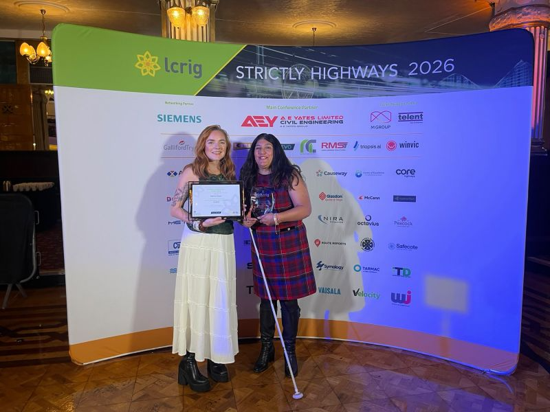Seema Flower, founder of Blind Ambition, proudly receives the Outstanding Contribution Award (Private Sector) at the Strictly Highways 2026 awards ceremony. Seema, wearing a red and navy tartan dress and holding her white cane, stands alongside the award presenter, who holds a framed certificate, in front of the event’s sponsor backdrop.